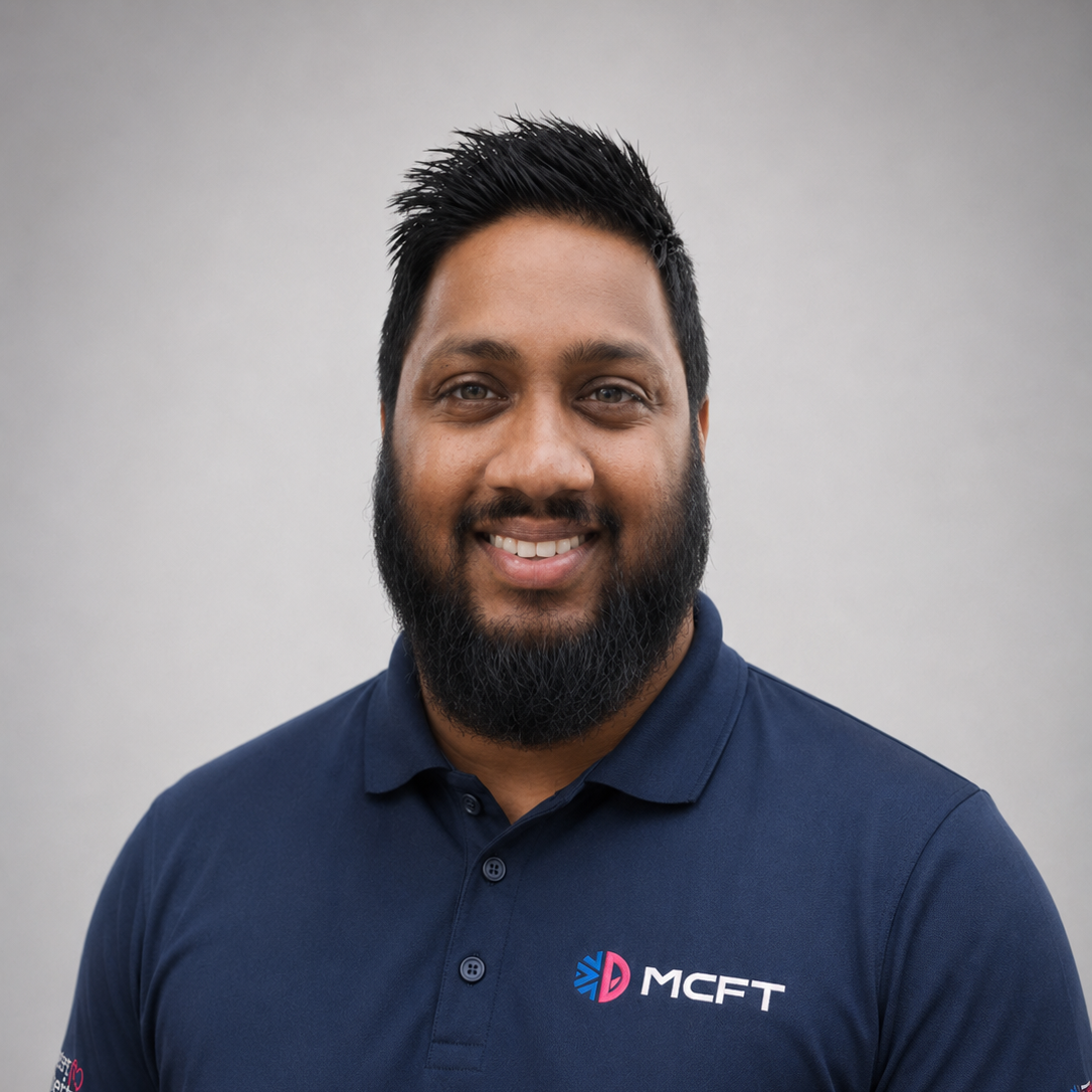Portrait of a man with dark hair and beard, wearing a navy blue polo shirt with an MCFT logo, smiling against a plain light gray background.