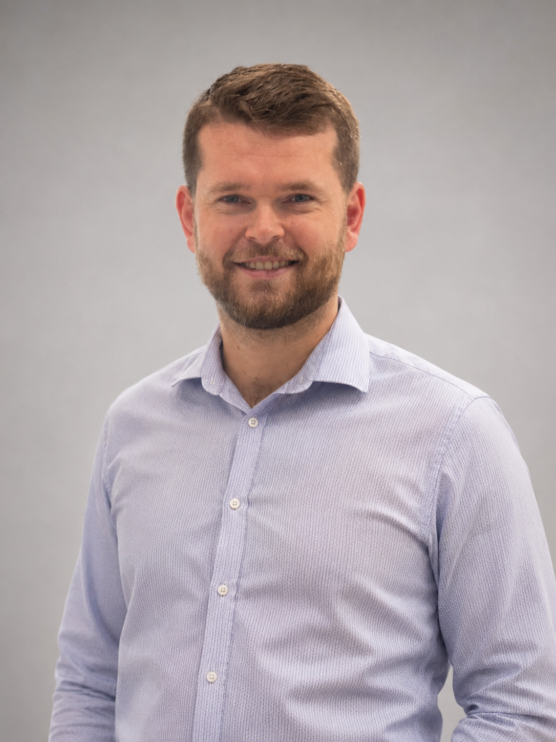 A smiling man with a beard and short brown hair wearing a light purple button-up shirt with a plain gray background.