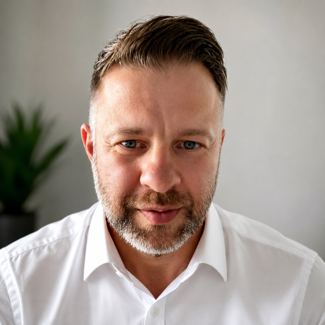 Close-up of a middle-aged man with blue eyes and a beard, wearing a white shirt, with a blurred green plant in the background.