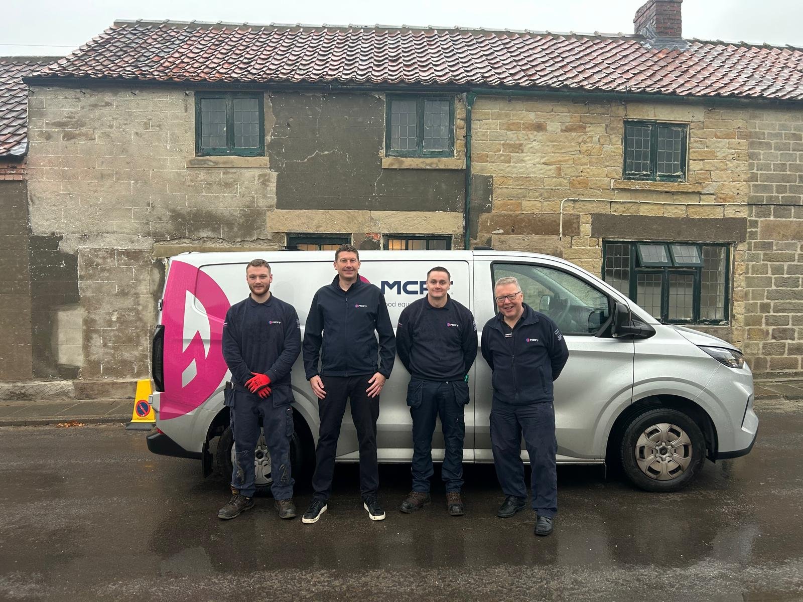 Four men standing in front of a white work van with a logo, in front of an old brick and stone building. They are dressed in work uniforms, and the man on the right is wearing glasses.