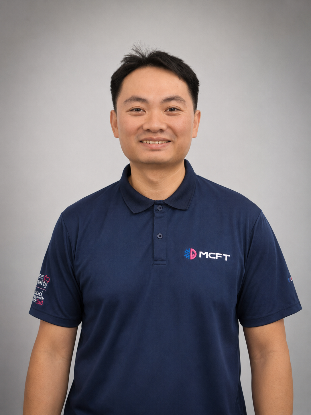 A man wearing a navy blue polo shirt with a company logo, smiling, against a gray background.