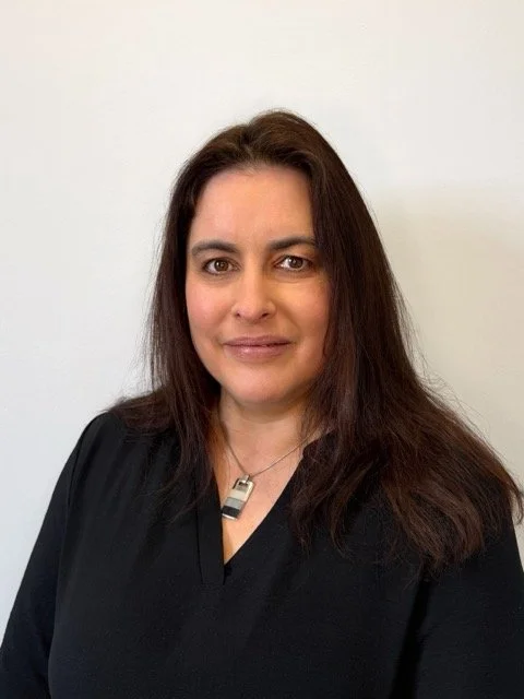 Portrait of a woman with long dark hair, wearing a black top and necklace, standing against a plain light-colored background.