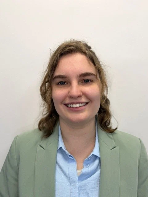 A young woman with wavy light brown hair smiling, wearing a light green blazer over a light blue button-up shirt, standing against a plain white wall.