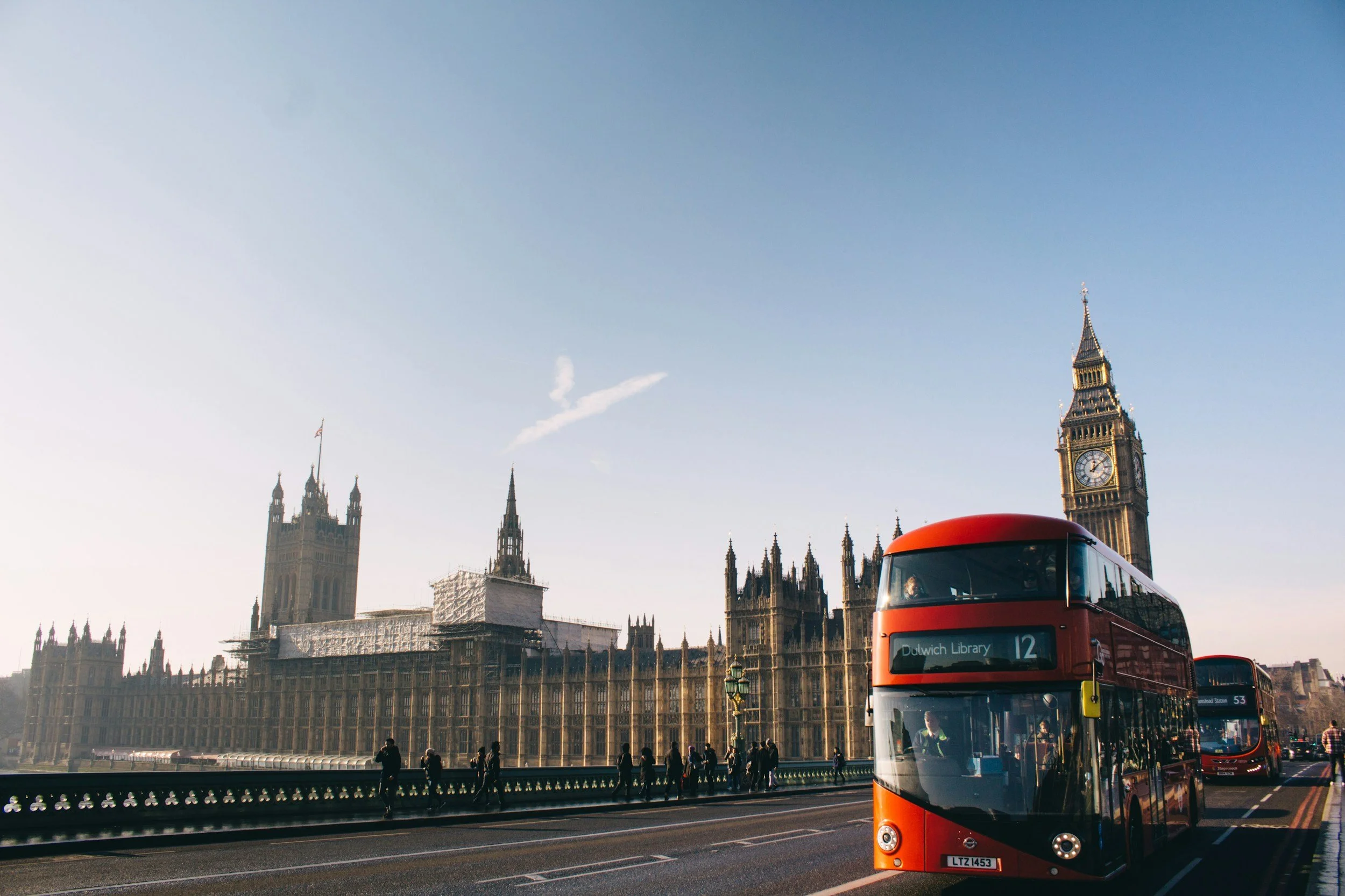 A red double-decker bus on the street in front of Big Ben and the Houses of Parliament in London, with pedestrians walking along the sidewalk in the late afternoon or early evening.