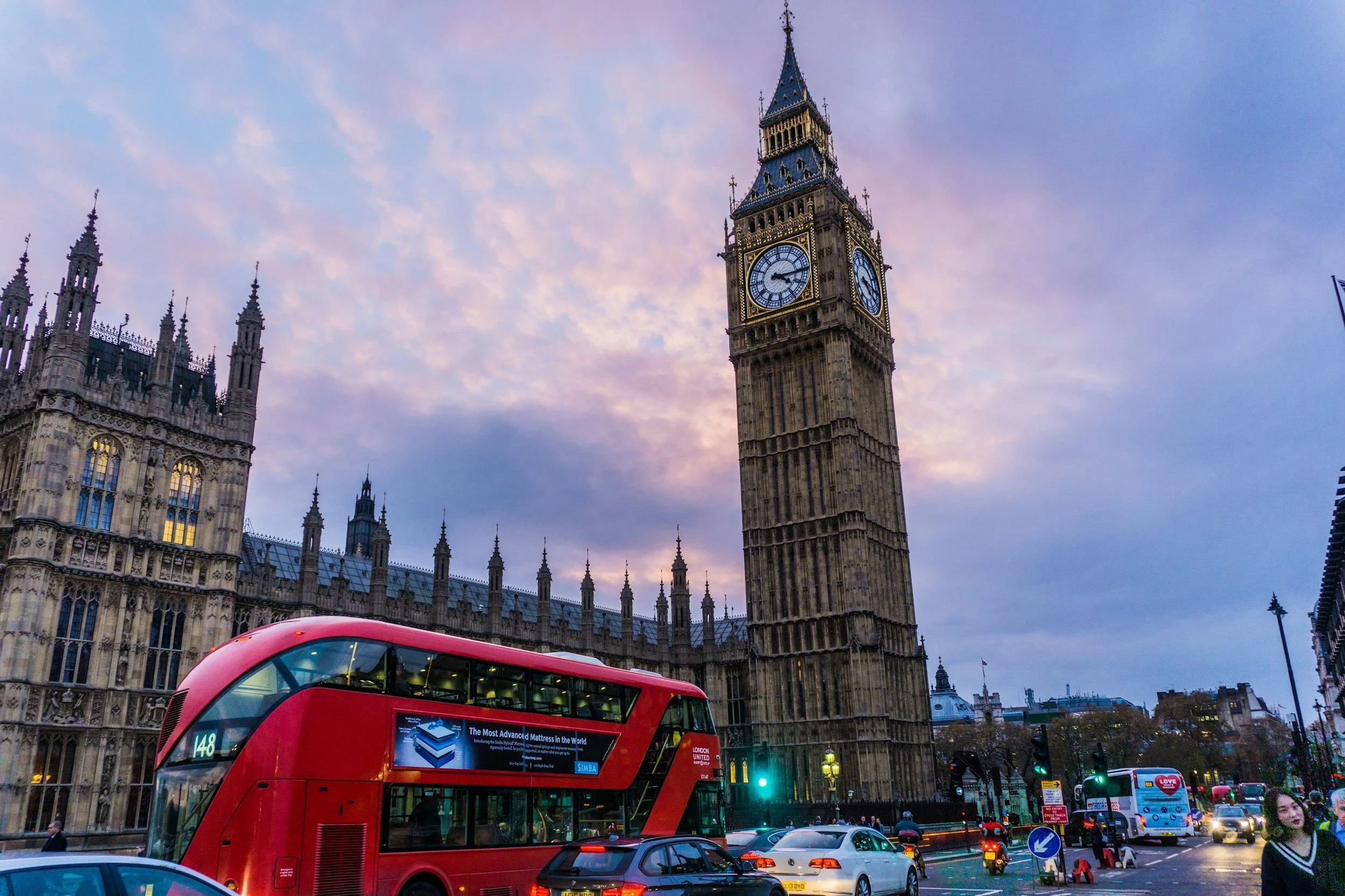 Big Ben clock tower in London with a red double-decker bus passing by and a cloudy sky at dusk.