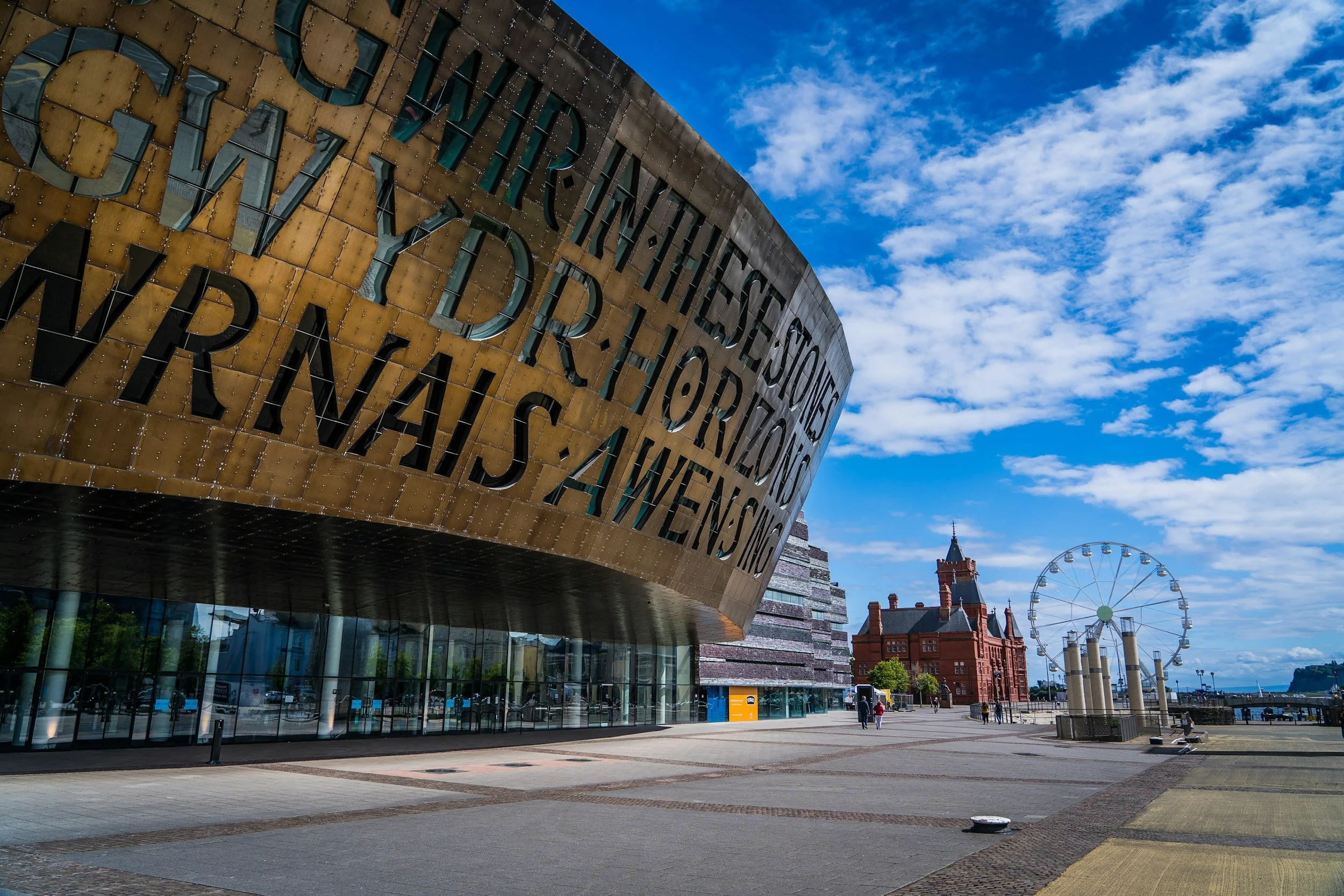 Exterior view of the Royal Liverpool Philharmonic Hall with modern architecture, large sign, glass entrance, a paved open area in front, and a clear sky with some clouds. There is a Ferris wheel and historic buildings in the background.