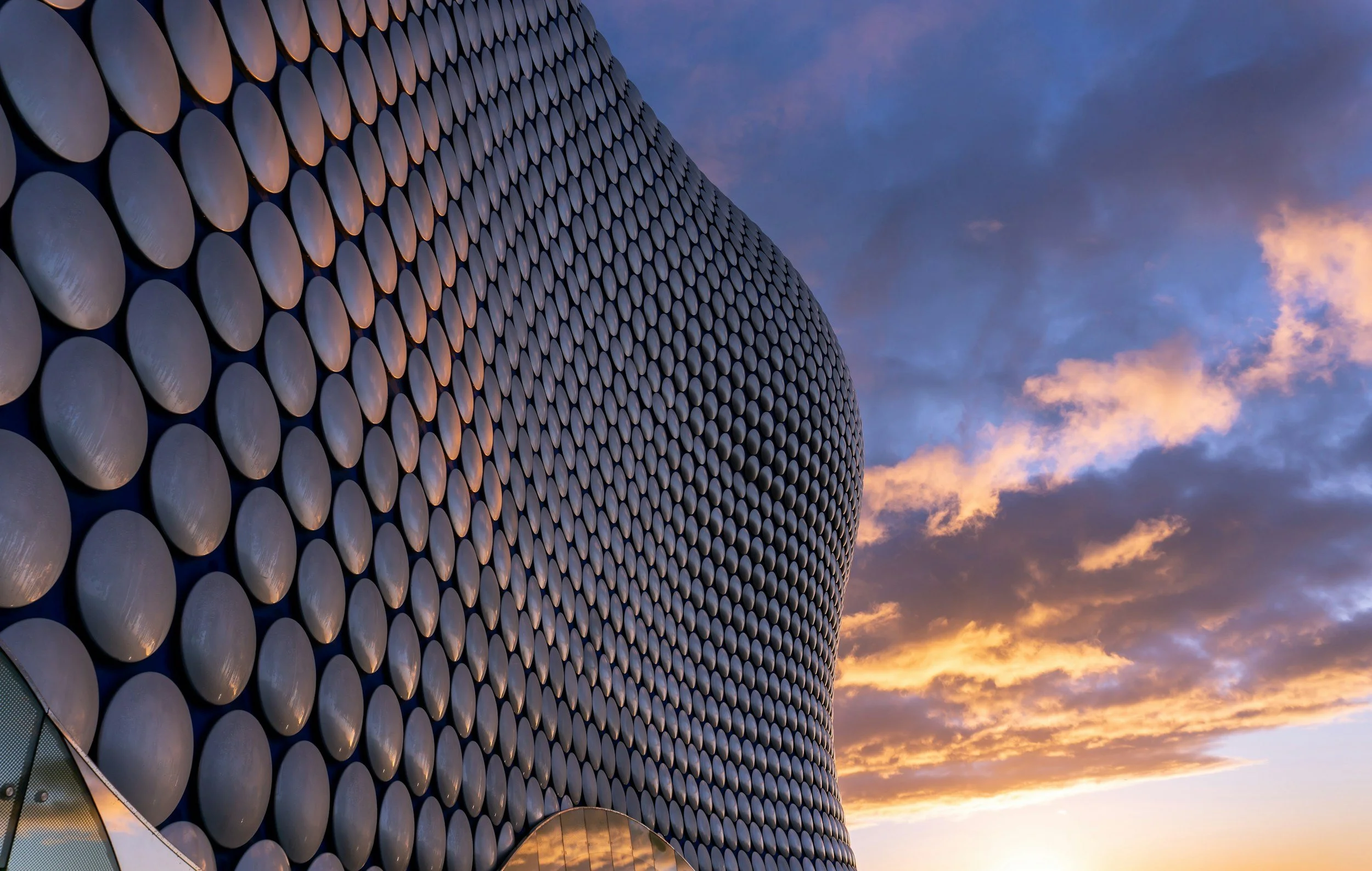 A modern building with a facade made up of numerous circular panels. The photo is taken during sunset or sunrise, with a sky showing dark clouds and patches of pink and orange hues.