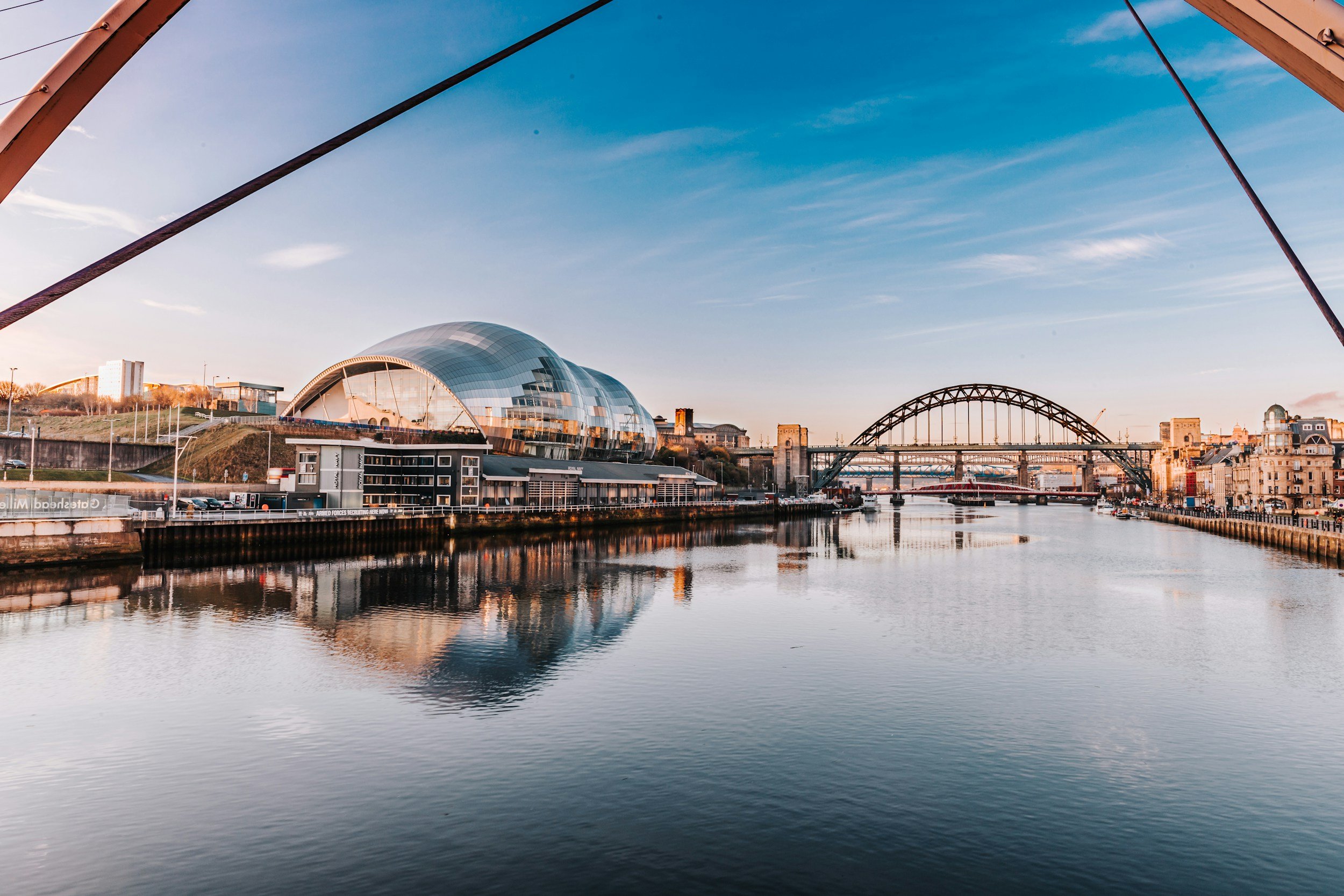 A cityscape with a modern, domed building and a bridge over a river, under a partly cloudy sky during sunset.