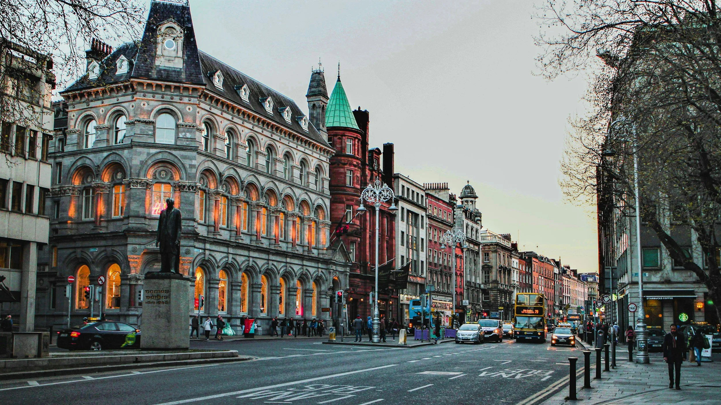 Street scene in a city with historic buildings, cars, buses, and pedestrians. There are ornate street lamps and a statue in the foreground. The sky is overcast, and the buildings feature various architectural styles, including a prominent building with arched windows and a statue of a man in front.