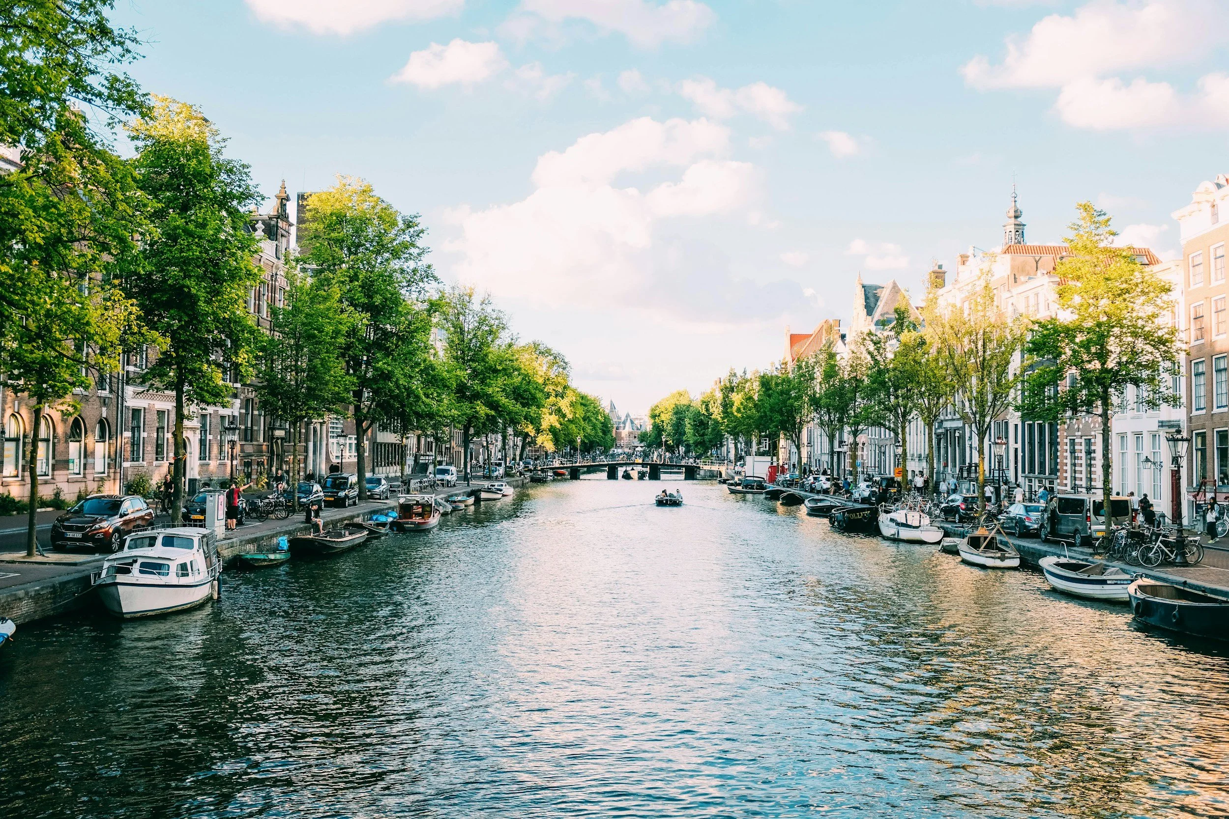 A scenic canal in a European city with boats docked along the sides, surrounded by trees and historic buildings under a partly cloudy sky.