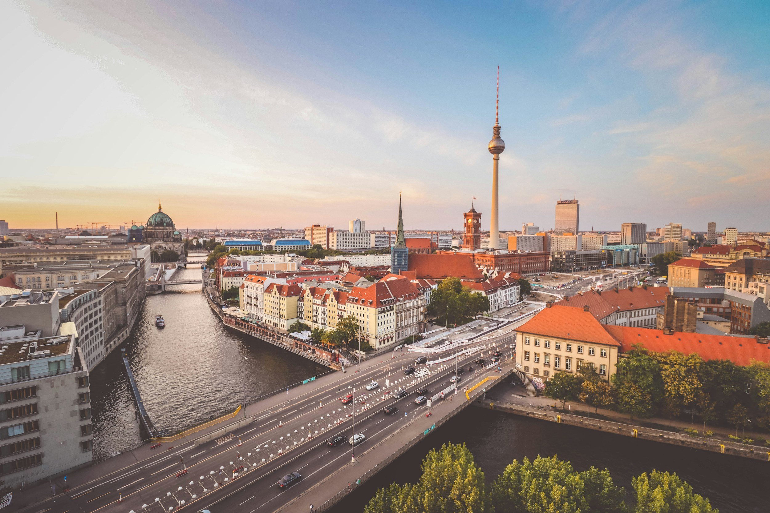 A city skyline during sunset featuring the Berlin TV Tower, a historic church with a green dome, and several modern buildings. A river cuts through the city with boats visible, and roads with cars run alongside the river and through the city. The sky is partly cloudy with warm tones from the setting sun.