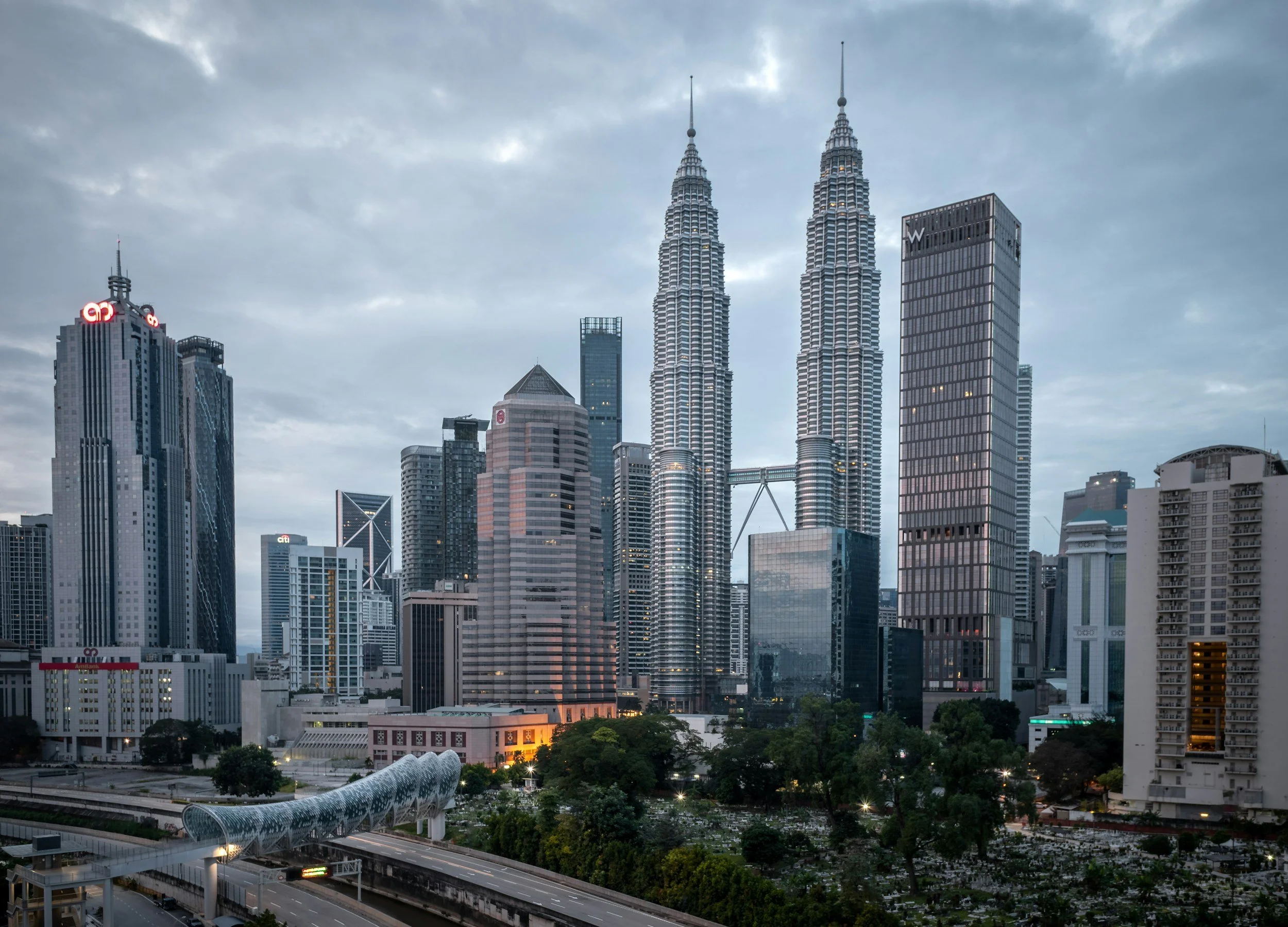 A city skyline featuring tall skyscrapers including the Petronas Towers, with a cloudy sky overhead. Some buildings have illuminated signs, and there is a bridge or walkway in the foreground with trees and a park area below.