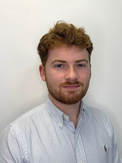 A young man with curly red hair and a beard, wearing a light blue striped button-up shirt, standing against a plain white wall.