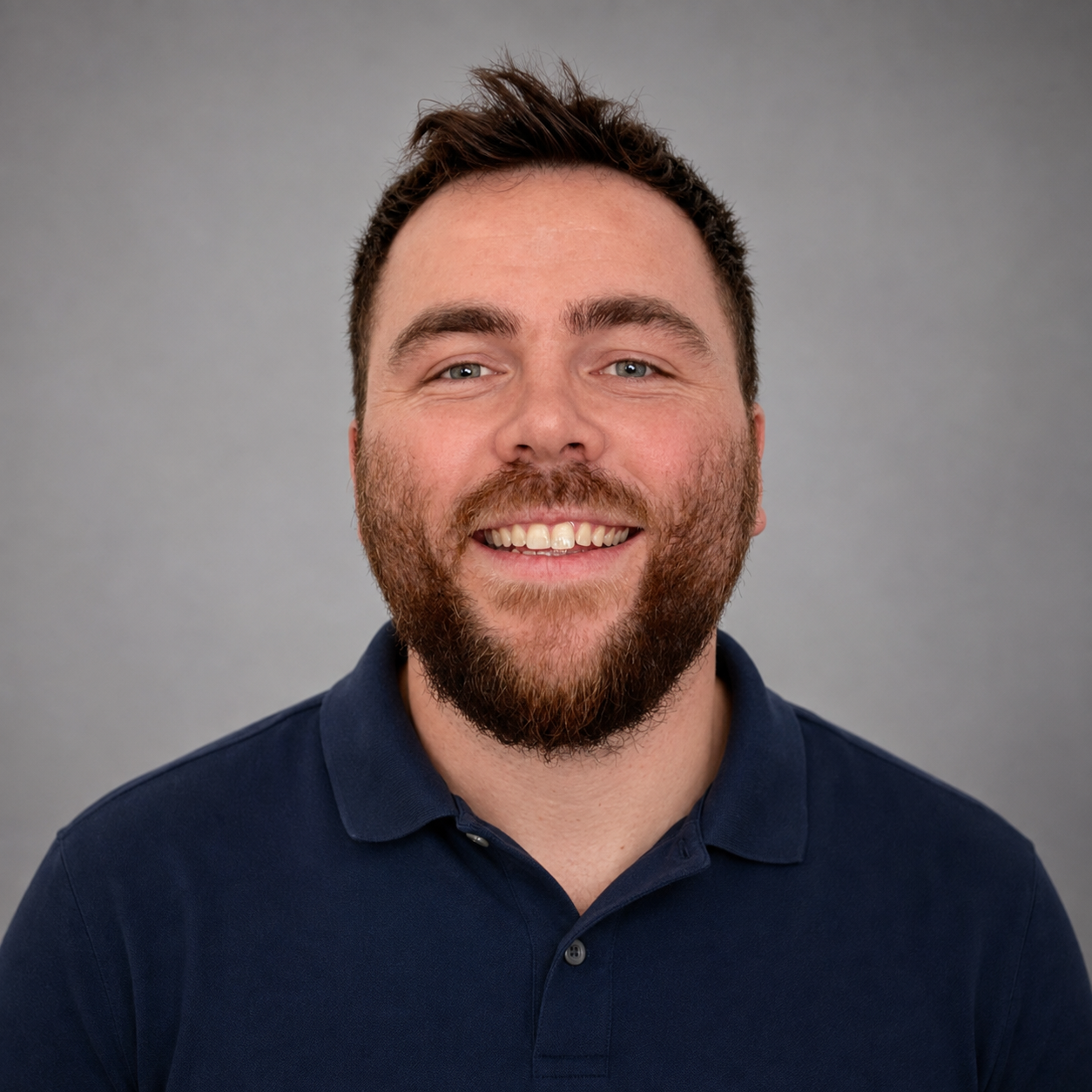 Headshot of a man with brown hair and a beard, smiling, wearing a navy blue polo shirt, against a gray background.