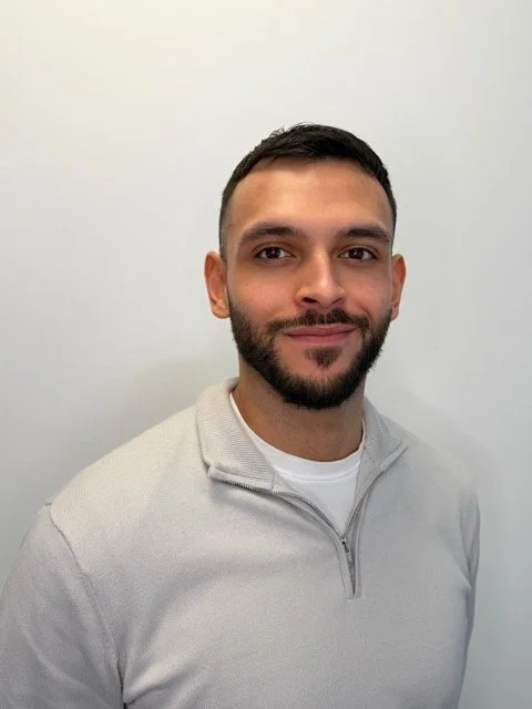 A young man with short dark hair, a beard, and light brown skin, smiling slightly, wearing a light-colored zip-up sweater, standing in front of a plain white wall.
