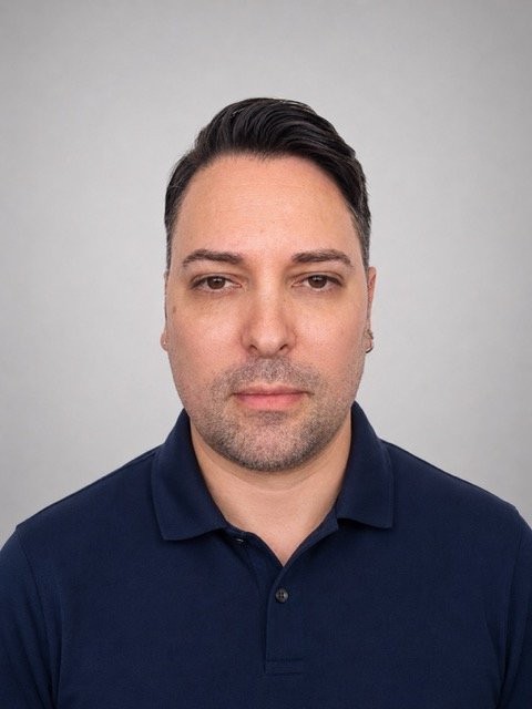 A portrait of a man with short dark hair, wearing a navy blue polo shirt, looking directly at the camera against a plain light gray background.