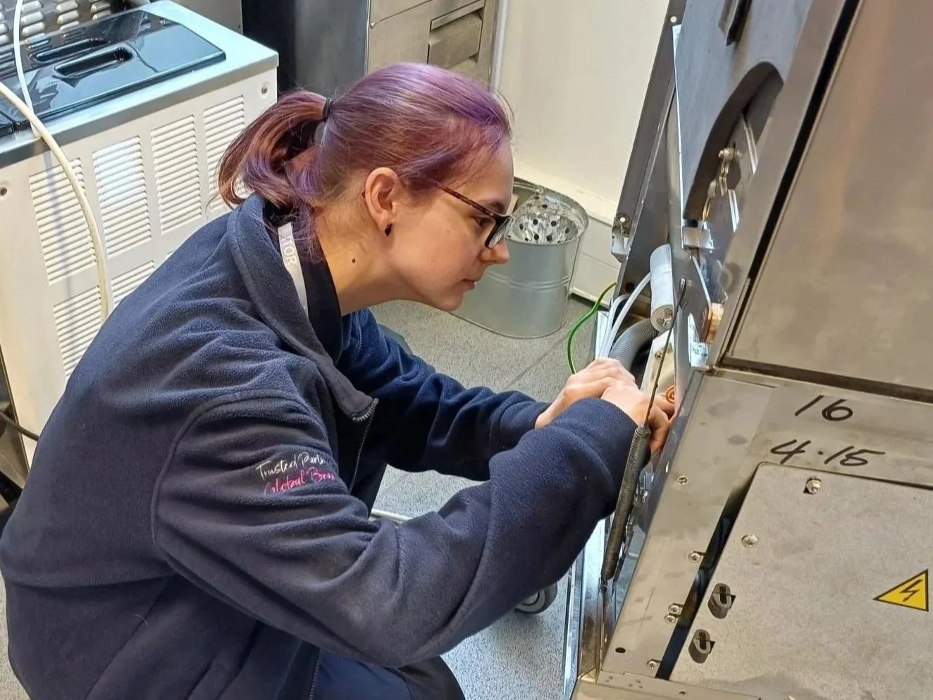 A woman with purple hair and glasses working on a laboratory machine in a lab. She is wearing a dark jacket with embroidered text.