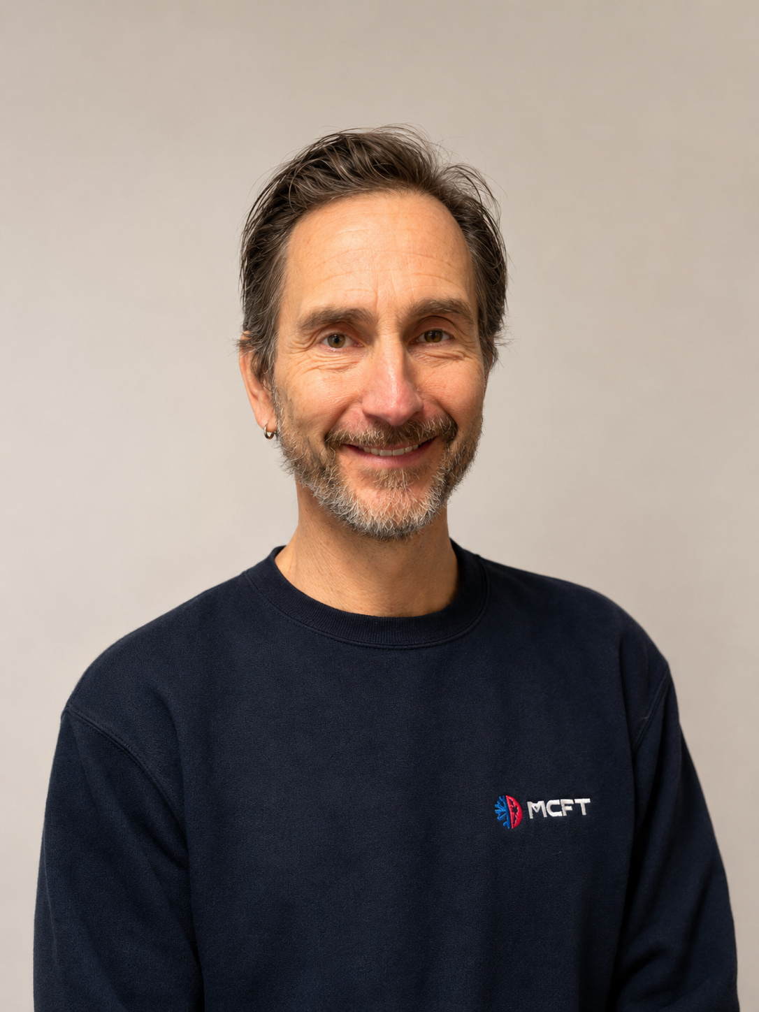 A man with short brown hair, a beard, and mustache, wearing a black sweatshirt with the 'MCFT' logo, smiling and standing against a plain gray background.