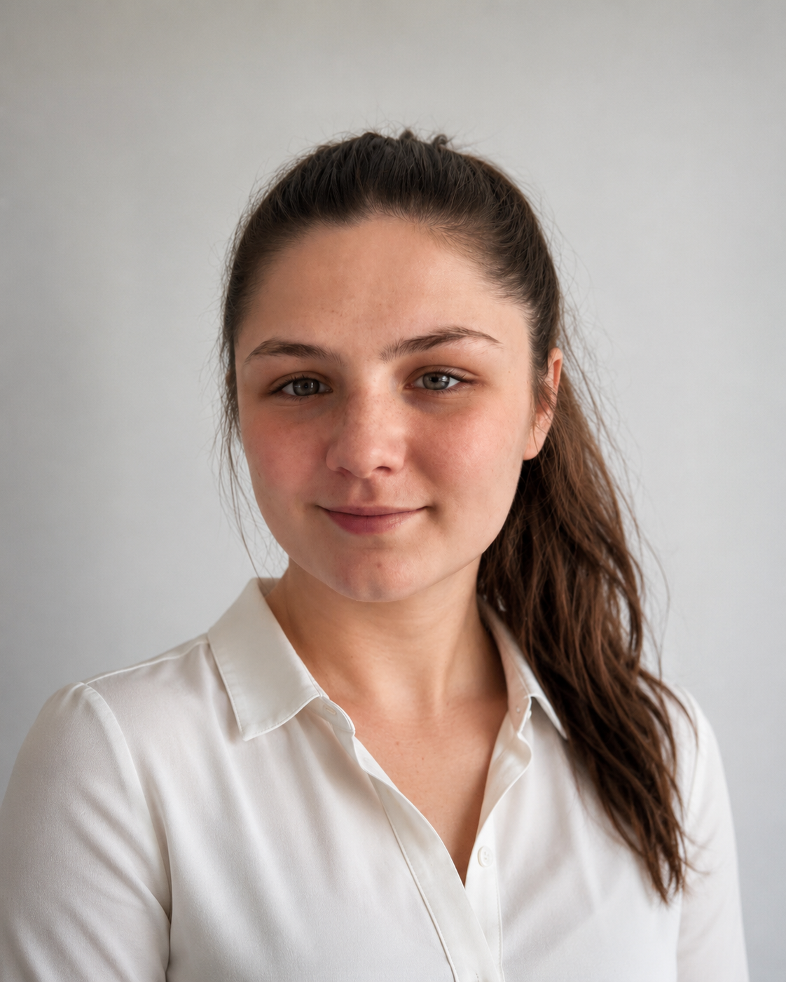Portrait of a young woman with long brown hair in a ponytail, wearing a white button-up shirt, smiling softly at the camera against a neutral gray background.