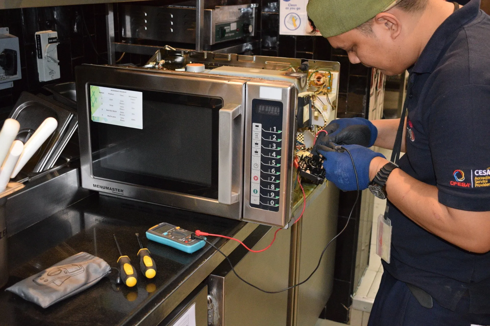 A person wearing gloves and a uniform working on repairing a microwave oven, using a multimeter probes and a screwdriver on a workbench in a kitchen or service area.