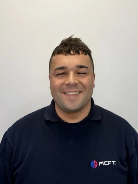 A smiling man with short dark hair, wearing a navy blue polo shirt with an 'MCFT' logo, standing against a plain light-colored wall.