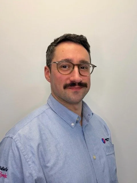 A man with glasses and a mustache wearing a light blue collared shirt with logos, posing against a plain light-colored background.