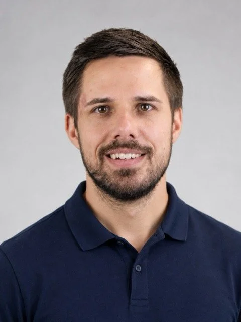 A man with brown hair and a beard, wearing a navy blue polo shirt, smiling at the camera against a gray background.