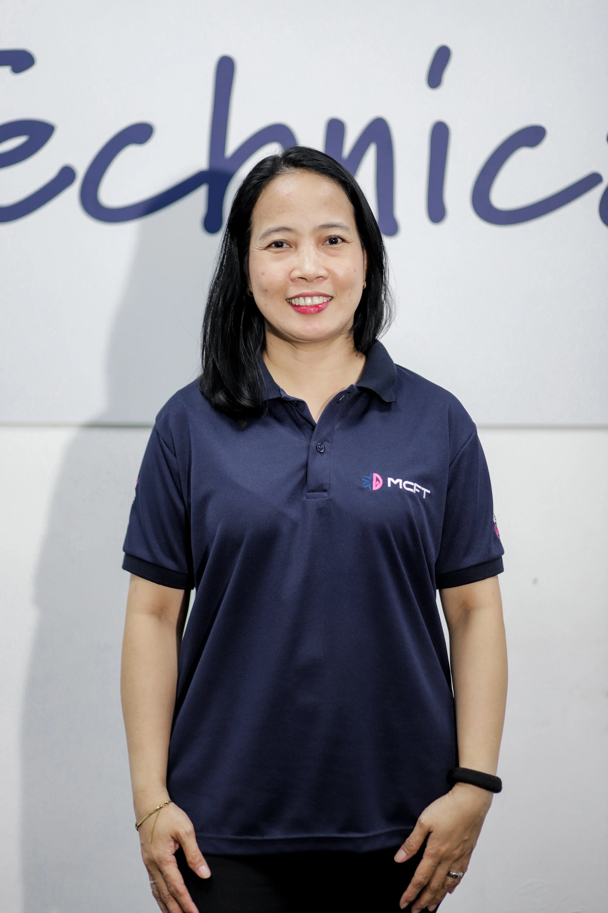 A woman with black hair wearing a navy blue polo shirt with a logo standing in front of a white wall with purple writing.