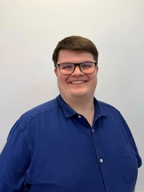 A young man with glasses and short brown hair, smiling, wearing a blue button-up shirt, standing against a white background.