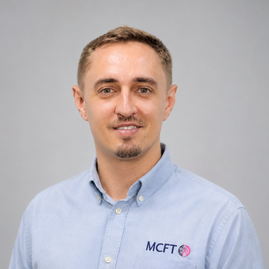 Headshot of young man with short light brown hair, goatee, wearing a light blue collared shirt with 'MCFT' logo on the chest, smiling slightly, against a plain gray background.