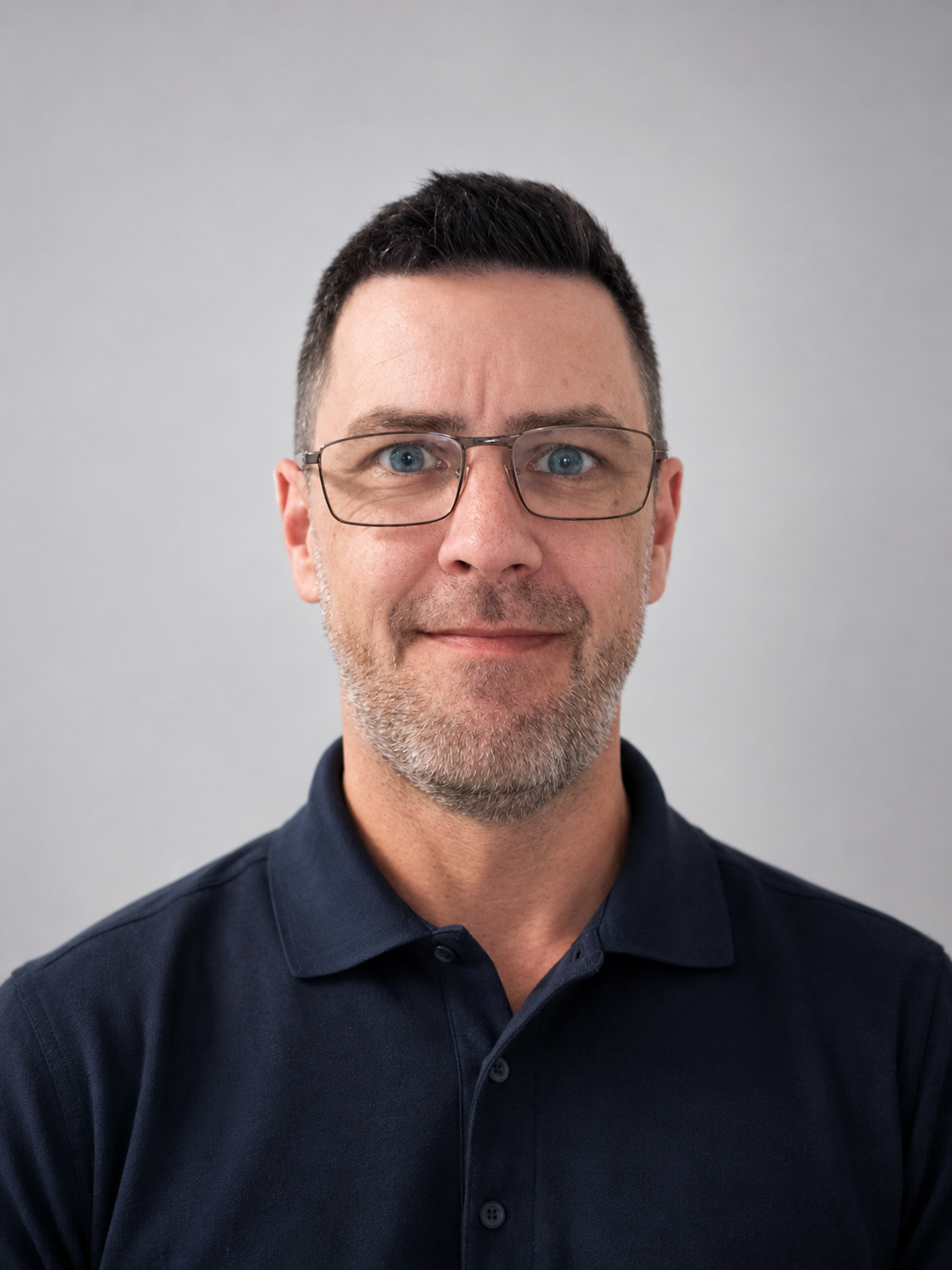 A middle-aged man with short dark hair and glasses, wearing a dark blue collared shirt, standing against a plain light gray background.