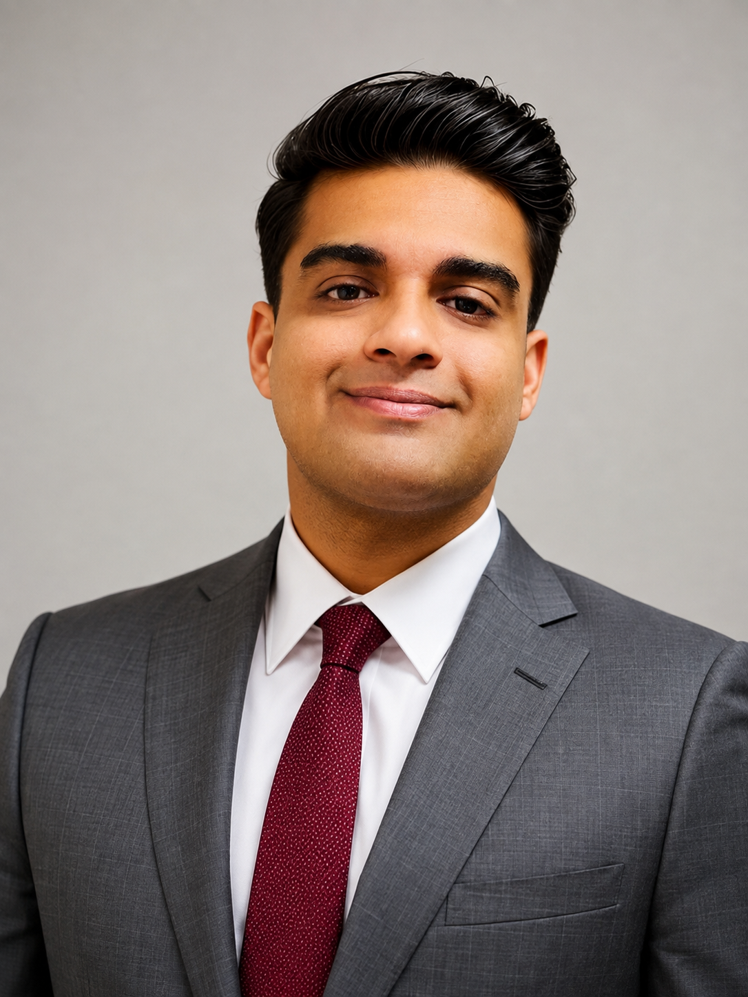 A young man with dark hair, wearing a gray suit, white shirt, and red tie, smiling at the camera against a plain gray background.