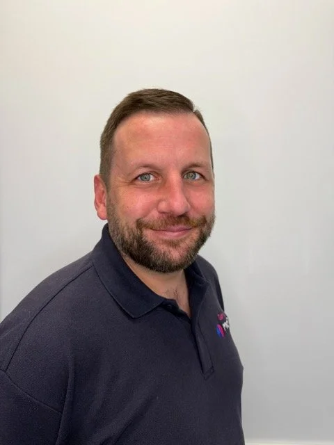 A man with short brown hair, a beard, and blue eyes, wearing a navy polo shirt with a logo on the chest, posing against a plain white wall.