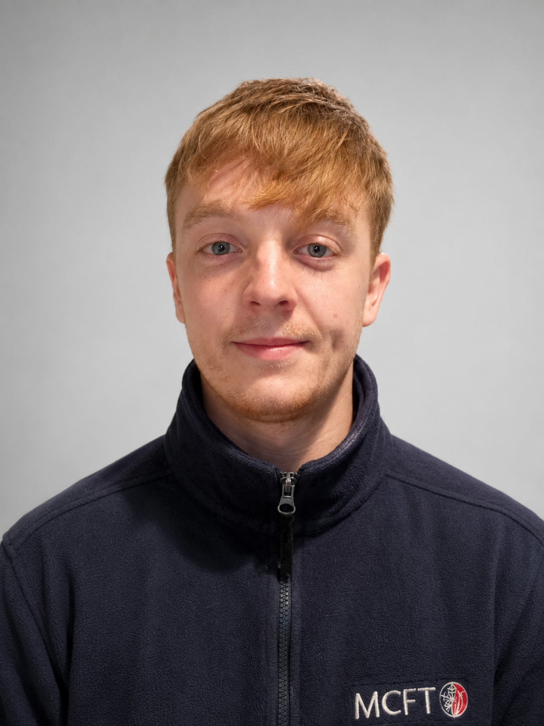 Young man with red hair, wearing a dark zip-up jacket with 'MCFT' logo, standing against a plain light gray background.