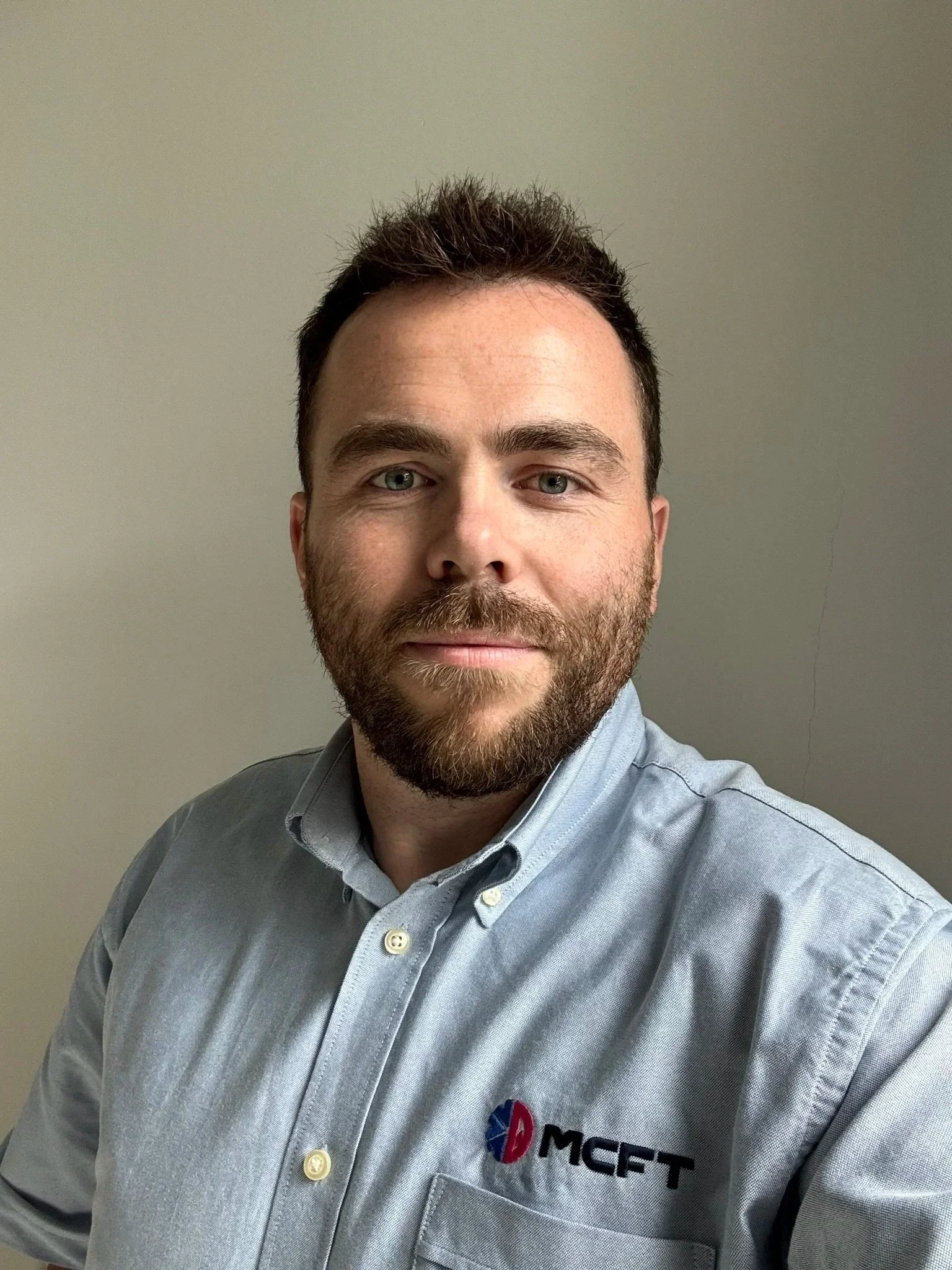 A man with short dark hair and a beard, wearing a gray button-up shirt with the logo 'MCFT' on it. He is smiling slightly and looking at the camera, standing against a plain gray wall.