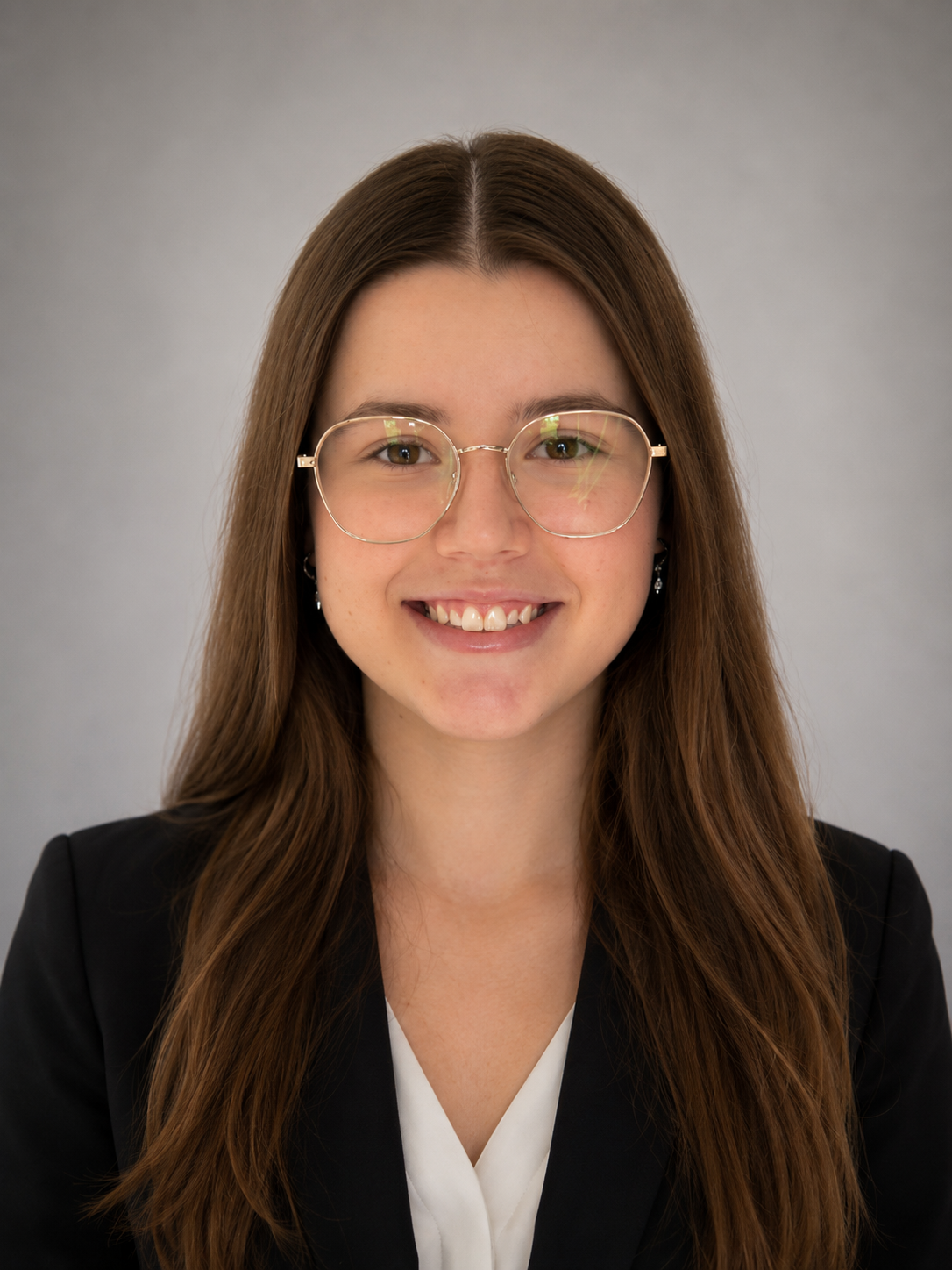 A young woman with long brown hair, glasses, and earrings, smiling in front of a plain gray background.