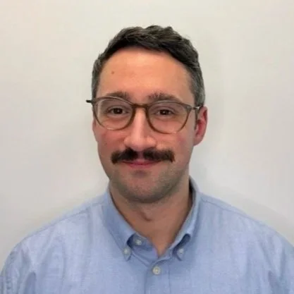A man with glasses, a mustache, and short dark hair, wearing a light blue button-up shirt, standing against a plain white background.