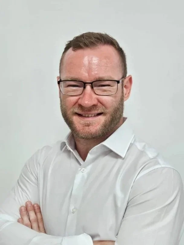 A man with glasses and a beard, wearing a white dress shirt, smiling with arms crossed, against a plain white background.