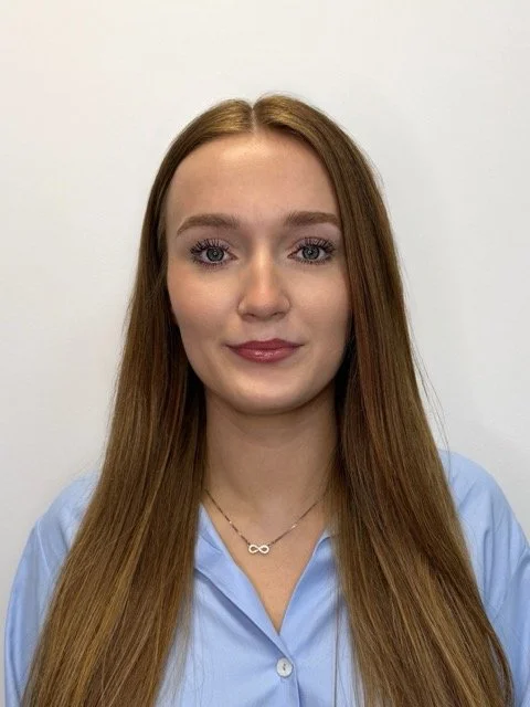 A young woman with long red hair, blue eyes, and fair skin wearing a light blue shirt and an infinity necklace, standing against a plain white background.