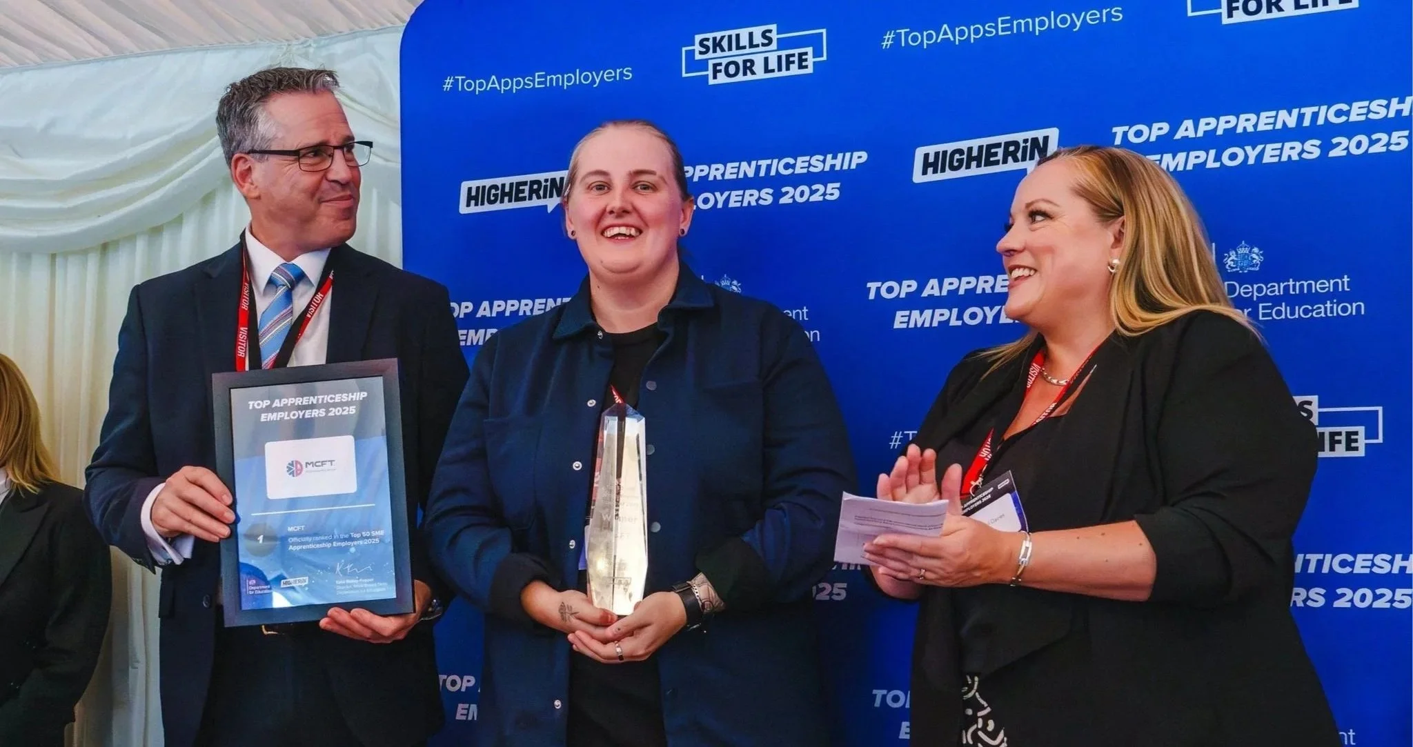 Three people at an award ceremony, with two of them holding awards and a framed certificate, standing in front of a blue backdrop with logos and text related to apprenticeships and employers.