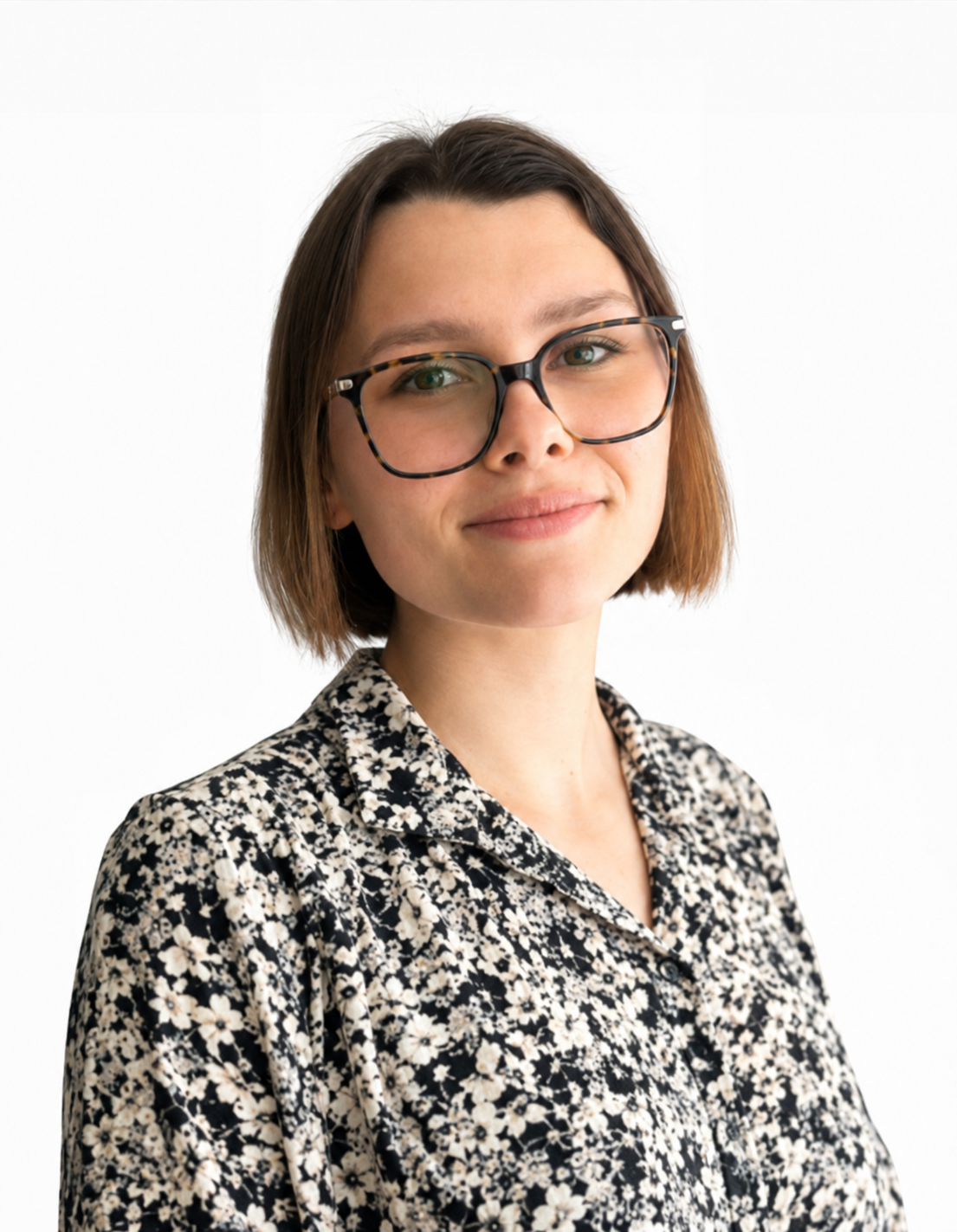 A woman with short brown hair and wearing large glasses, smiling softly, dressed in a floral patterned shirt, against a plain white background.