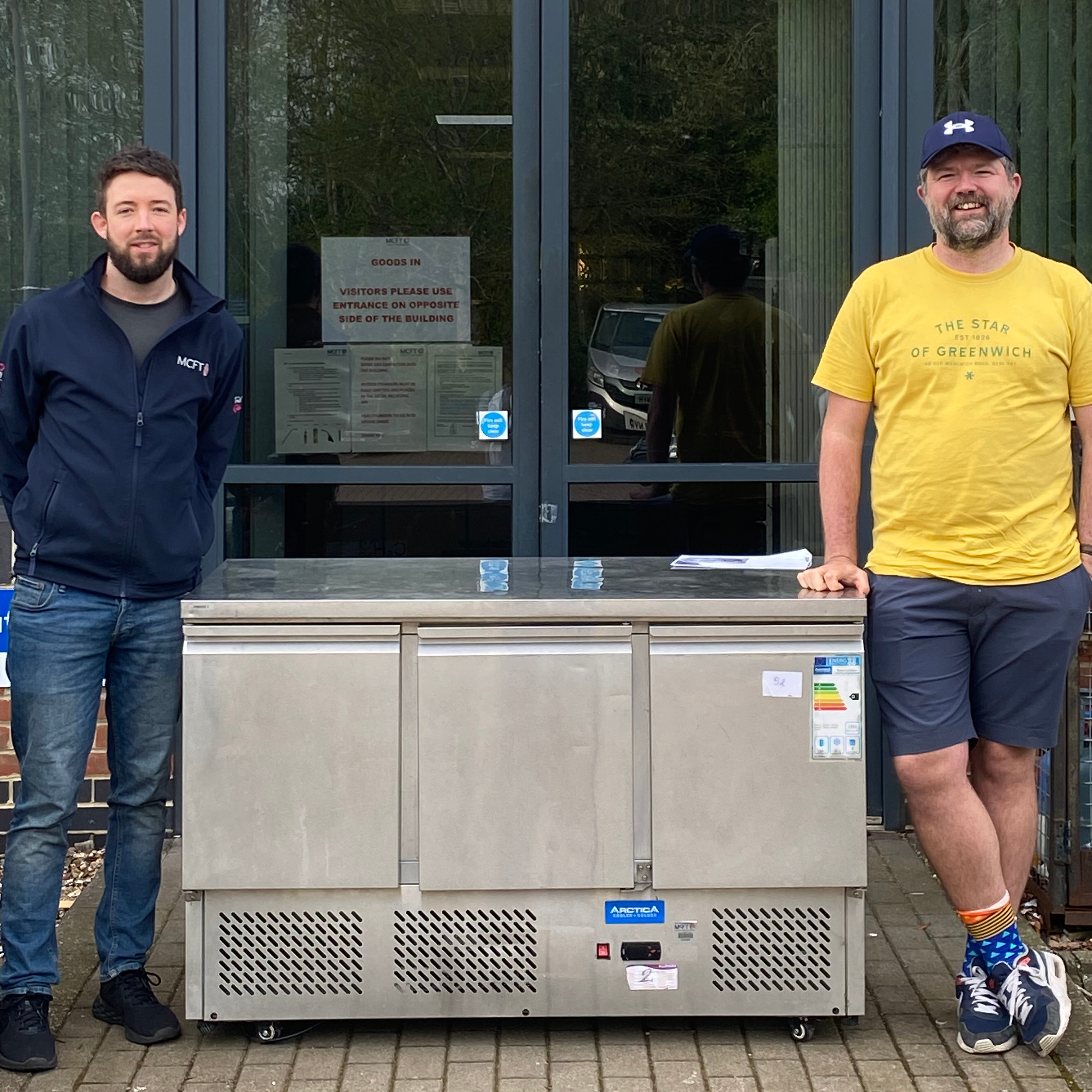 Two men standing outside, one on the left wearing a dark jacket and jeans, the other on the right in a yellow T-shirt, shorts, and a cap. They are positioned beside a large industrial ice machine with a sign on the glass door behind them. The background includes reflective glass and some outdoor elements.
