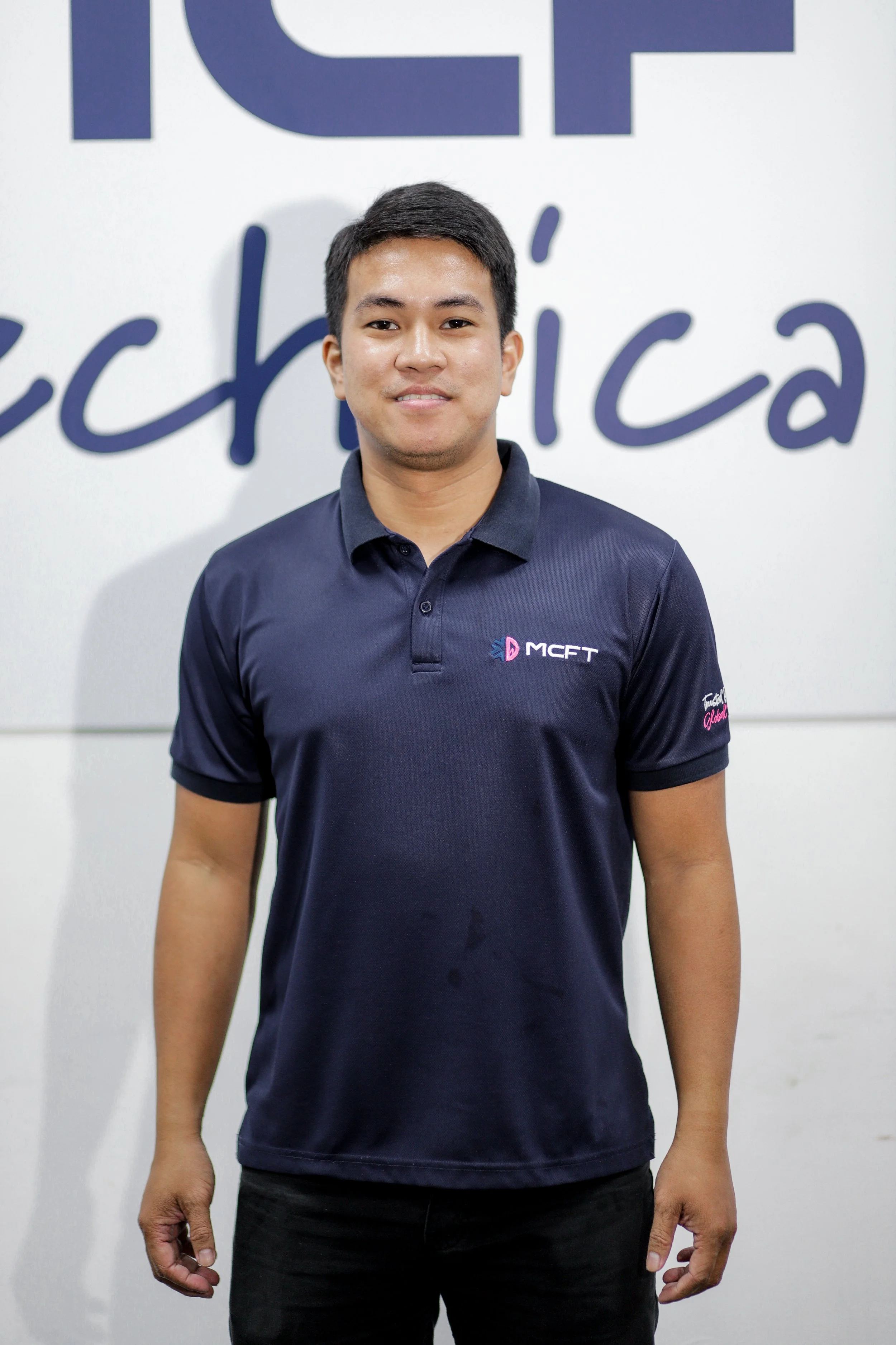 A young man standing in front of a wall with the partial text 'ICT' and 'Edh'. He is wearing a navy blue polo shirt with the logo 'MCFT' and 'Trust' on the sleeve. He is smiling subtly and has short black hair.