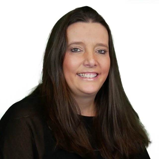 A woman with long brown hair smiling, wearing a black top, against a white background.