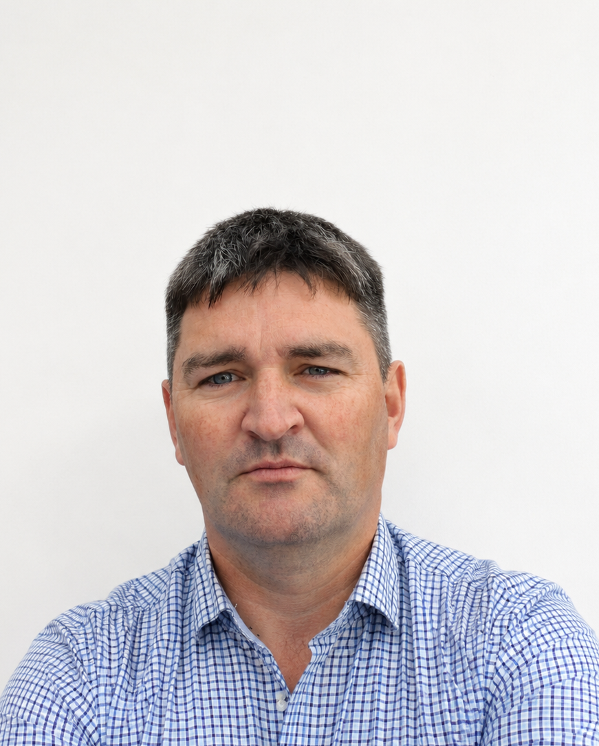 A middle-aged man with short, dark hair and light skin, wearing a blue checkered shirt, looking directly at the camera against a plain white background.