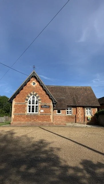 A small brick church with a pointed roof, arched window, and a sign that reads "Village Hall," under a clear blue sky.