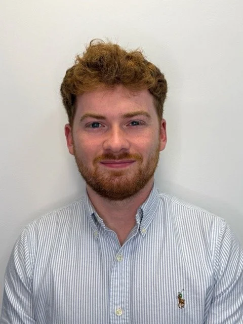 Headshot of a young man with red curly hair and a beard, wearing a light blue and white striped button-up shirt with a small logo on the chest, smiling in front of a plain white background.
