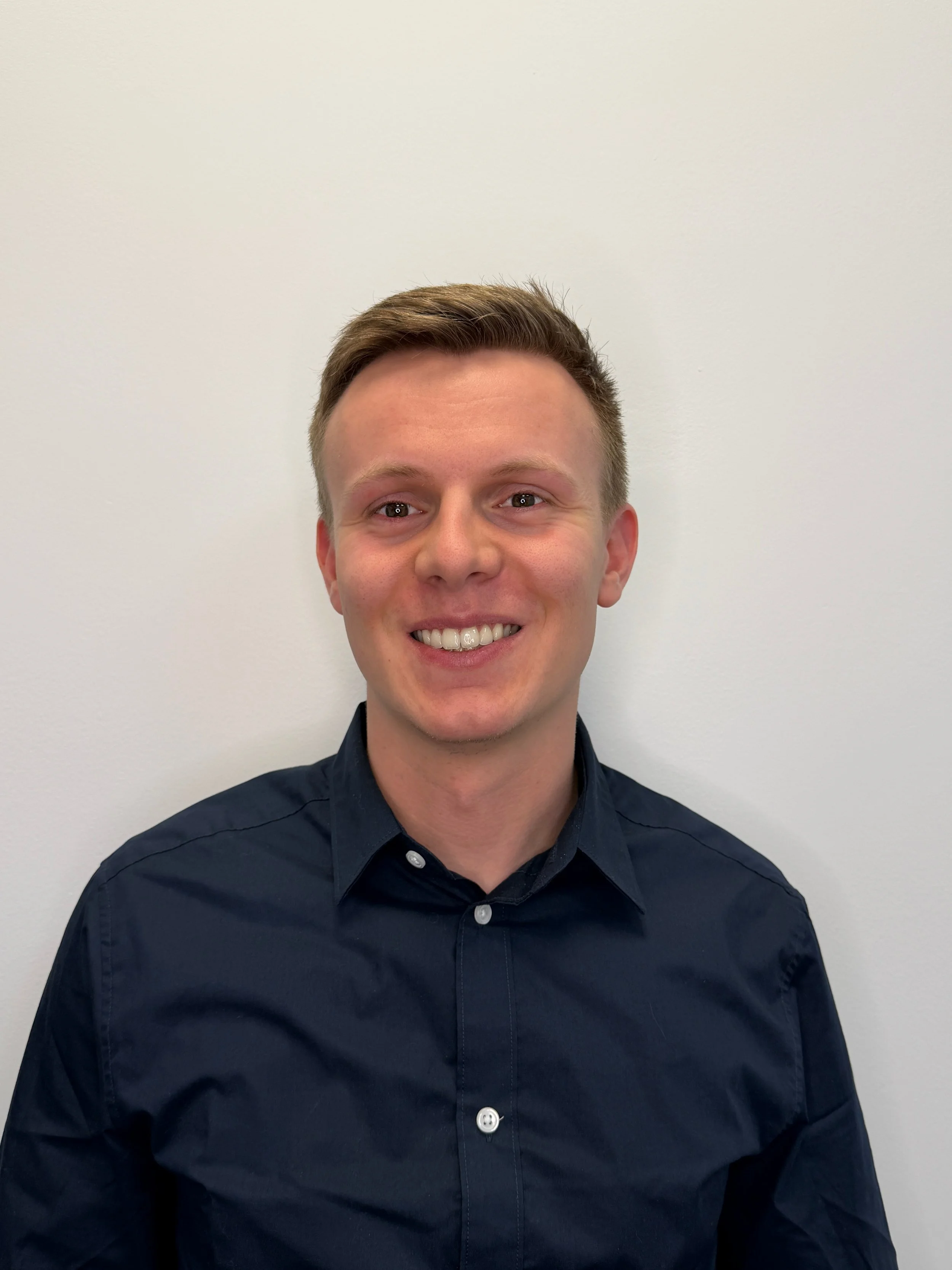 Portrait of a smiling young man with short brown hair, wearing a dark blue button-up shirt, standing against a plain white wall.