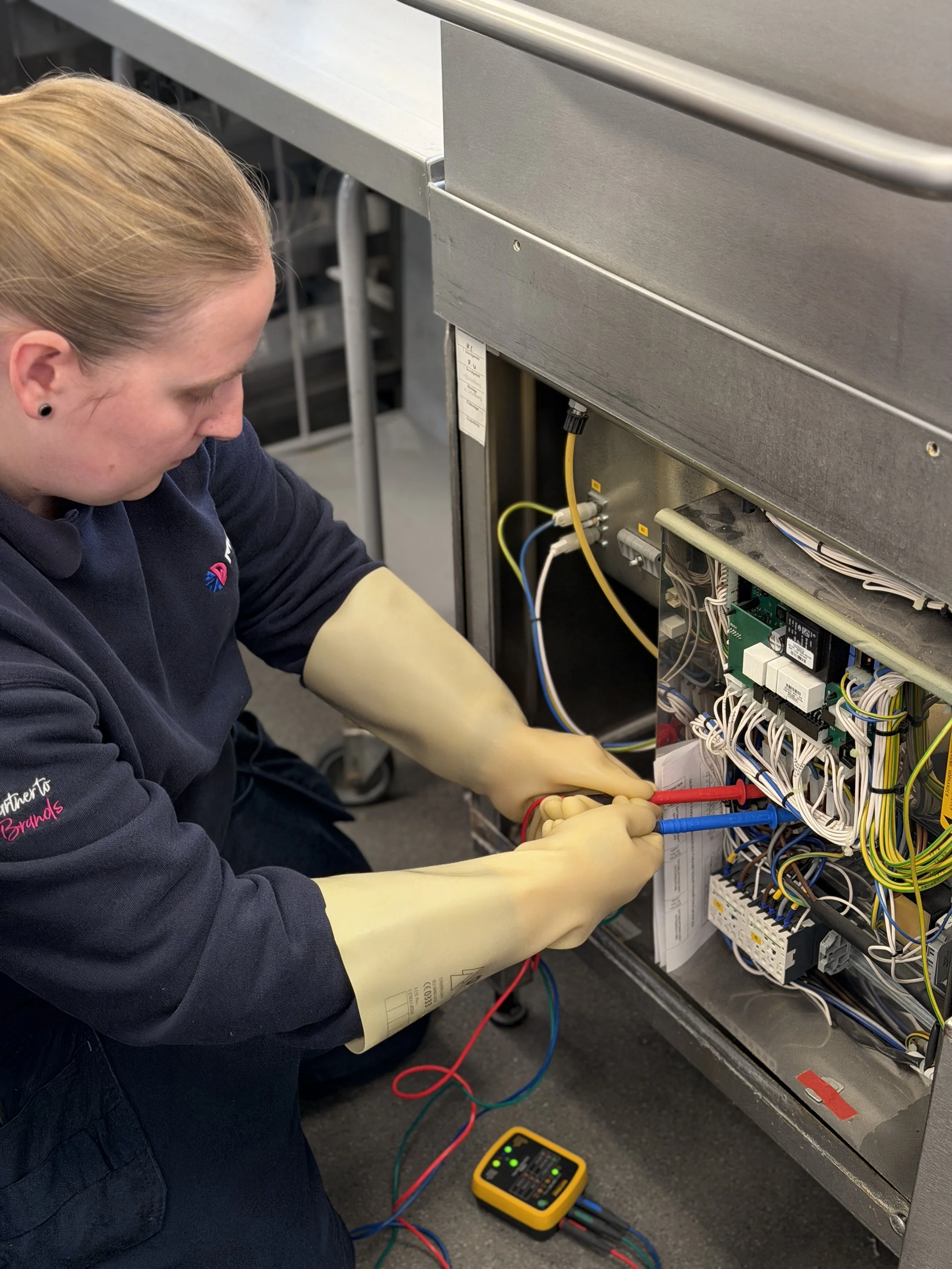 A technician wearing yellow gloves is working on the internal wiring of an electrical panel or control box, using a multimeter to test connections.
