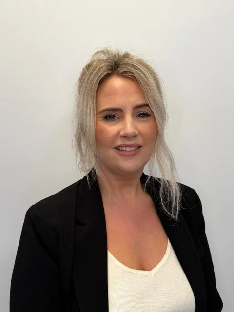 A woman with blonde hair in a loose updo, wearing a black blazer and a cream-colored top, standing against a plain white background.
